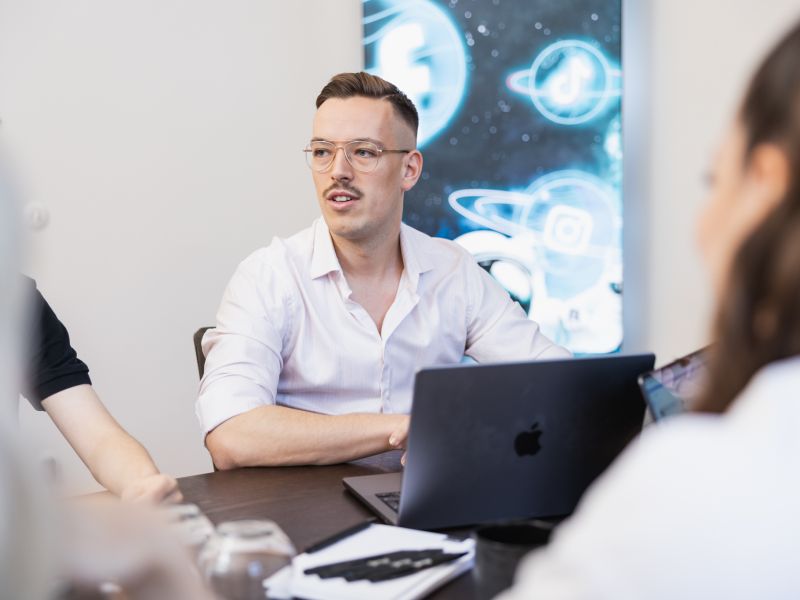 Ein Mann mit Brille sitzt an einem Tisch und diskutiert. Vor ihm steht ein aufgeklappter Laptop. An der Wand im Hintergrund sind Grafiken von Planeten und Social-Media-Symbolen zu sehen. Im Vordergrund sind zwei Personen teilweise zu sehen.
